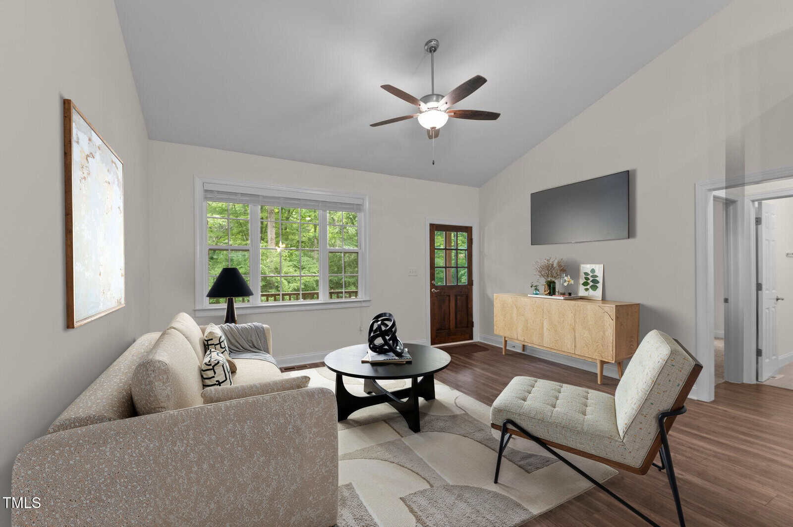 A modern living room with a beige sofa, patterned chair, round coffee table, light wood floors, sideboard, large wall art, and a ceiling fan. Large windows and a glass door reveal green trees outside.