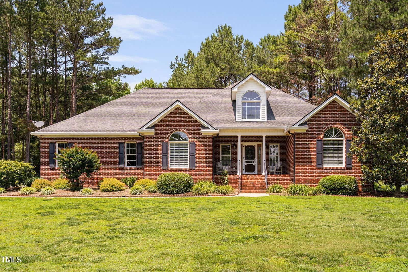 Single-story brick house with three arched front windows, a central door with sidelights, a gabled roof with a dormer window, and landscaped shrubs in front. Surrounded by green lawn and trees.