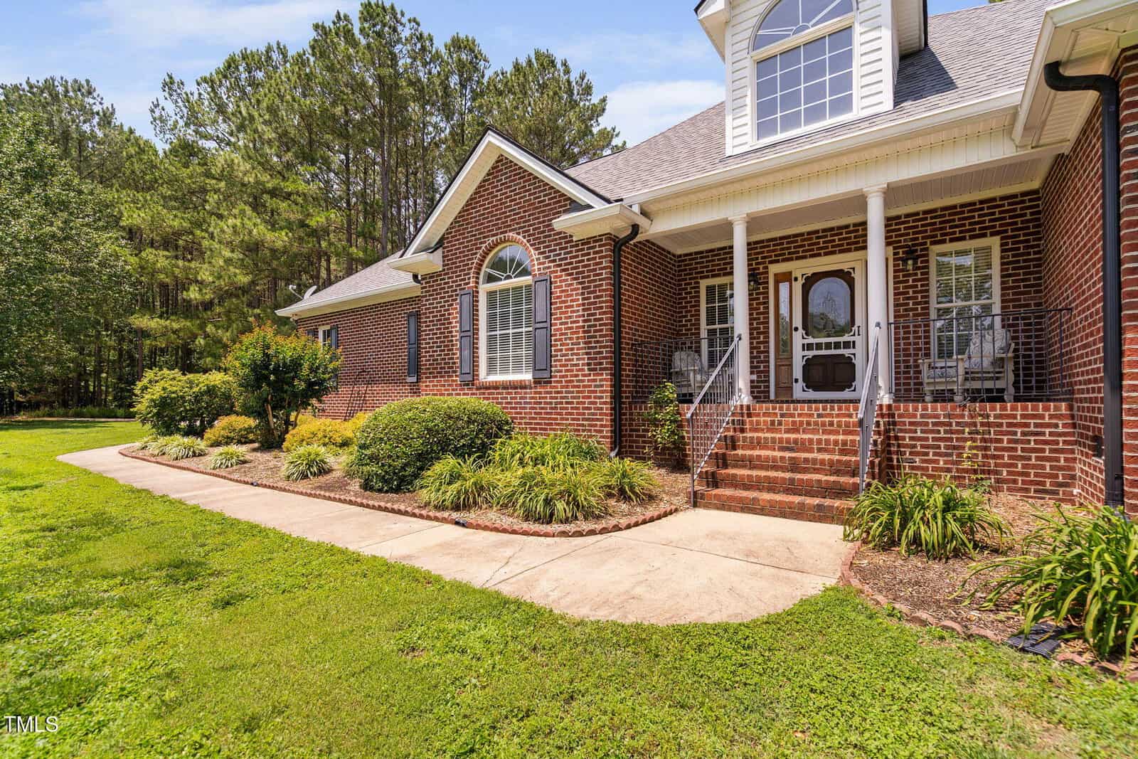 Brick house with white trim, front porch, black railings, and arched window. Concrete walkway curves past landscaping with shrubs and grass. Tall trees line the background under a blue sky.