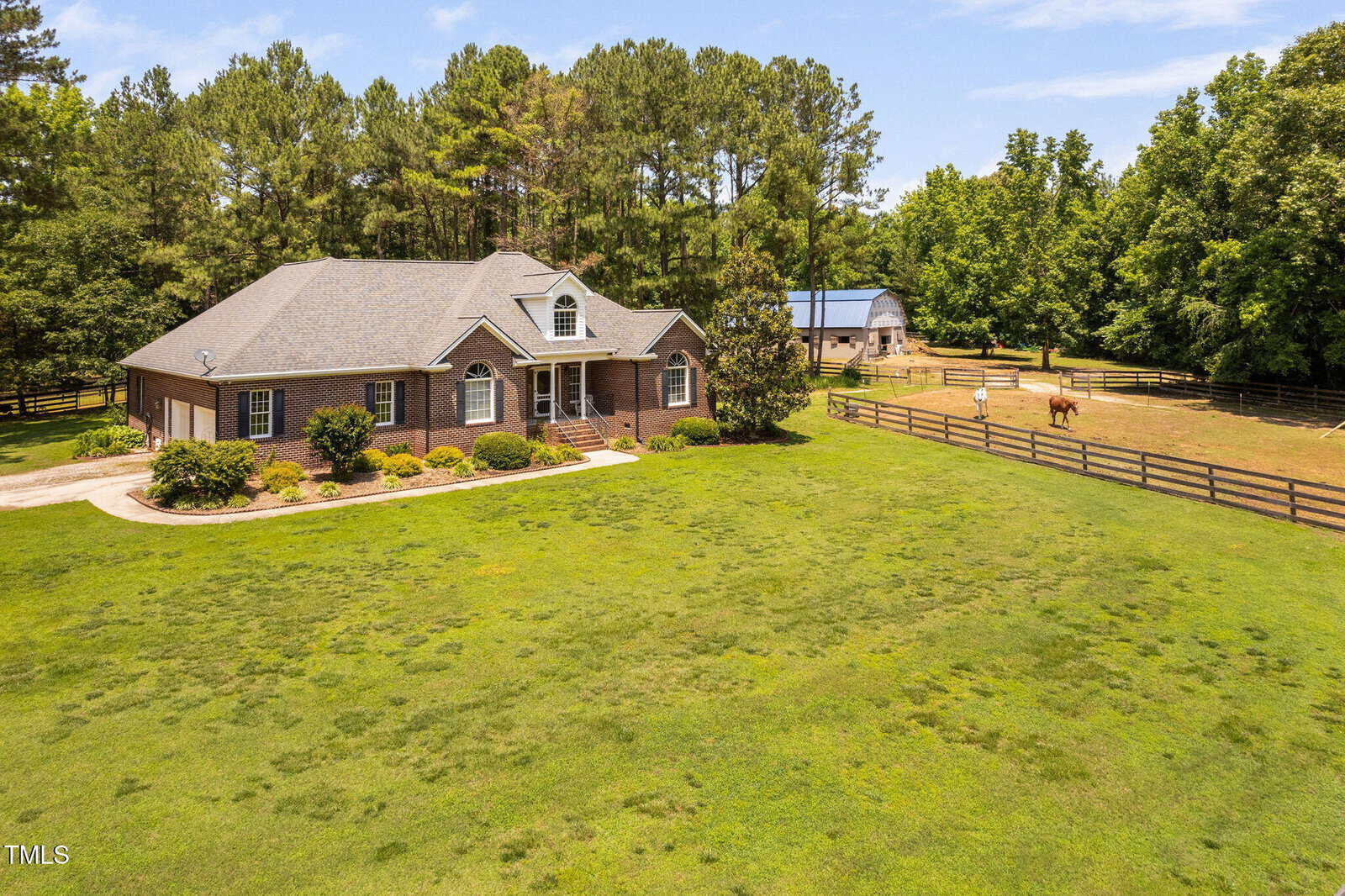 A brick house with a gray roof sits on a large grassy lot, bordered by a wooden fence. There is a barn and a horse in a fenced area, with trees and blue sky in the background.