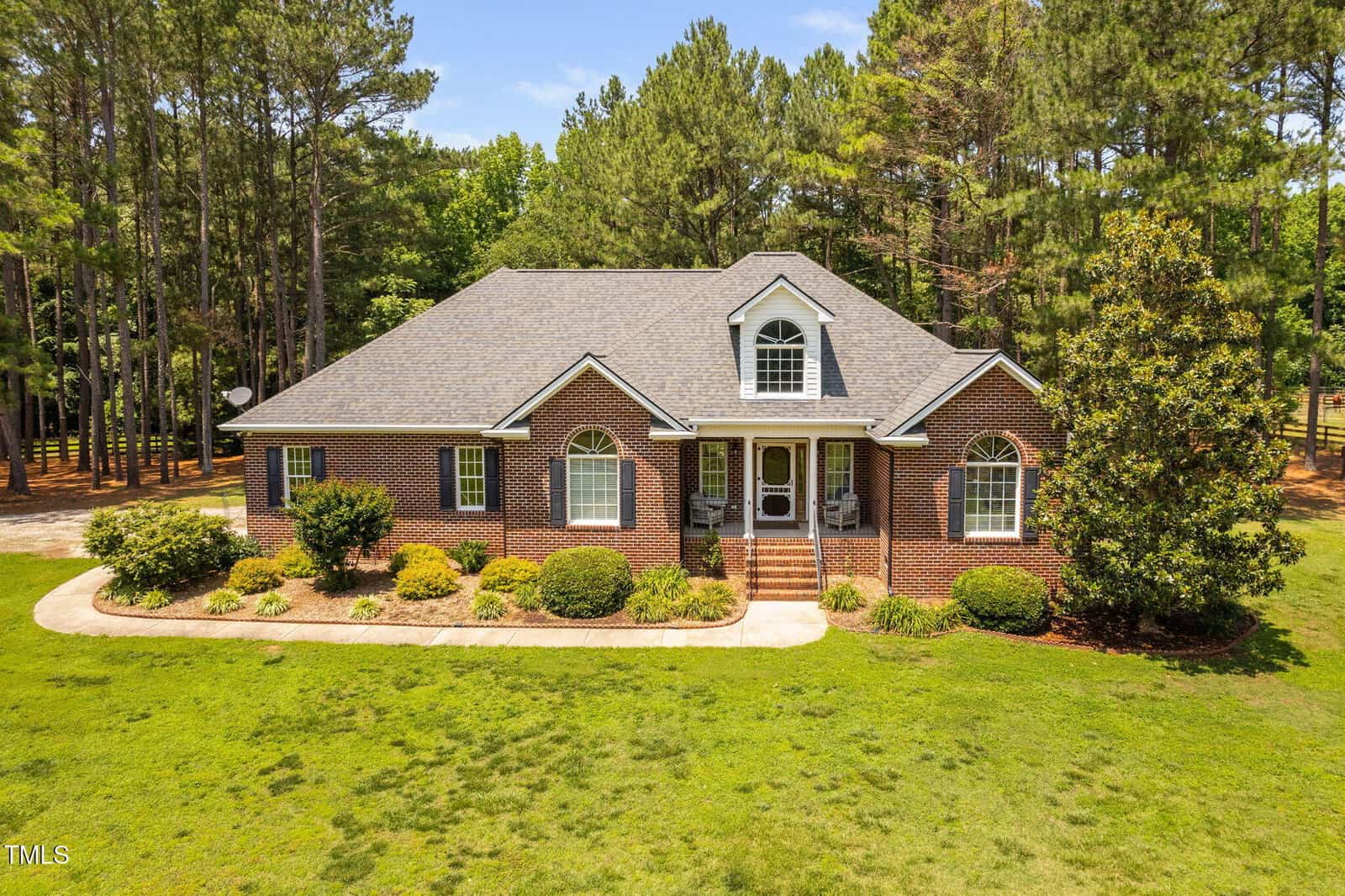Single-story brick house with arched windows, a dormer, and front steps. Surrounded by grass, shrubs, and tall pine trees under a blue sky.