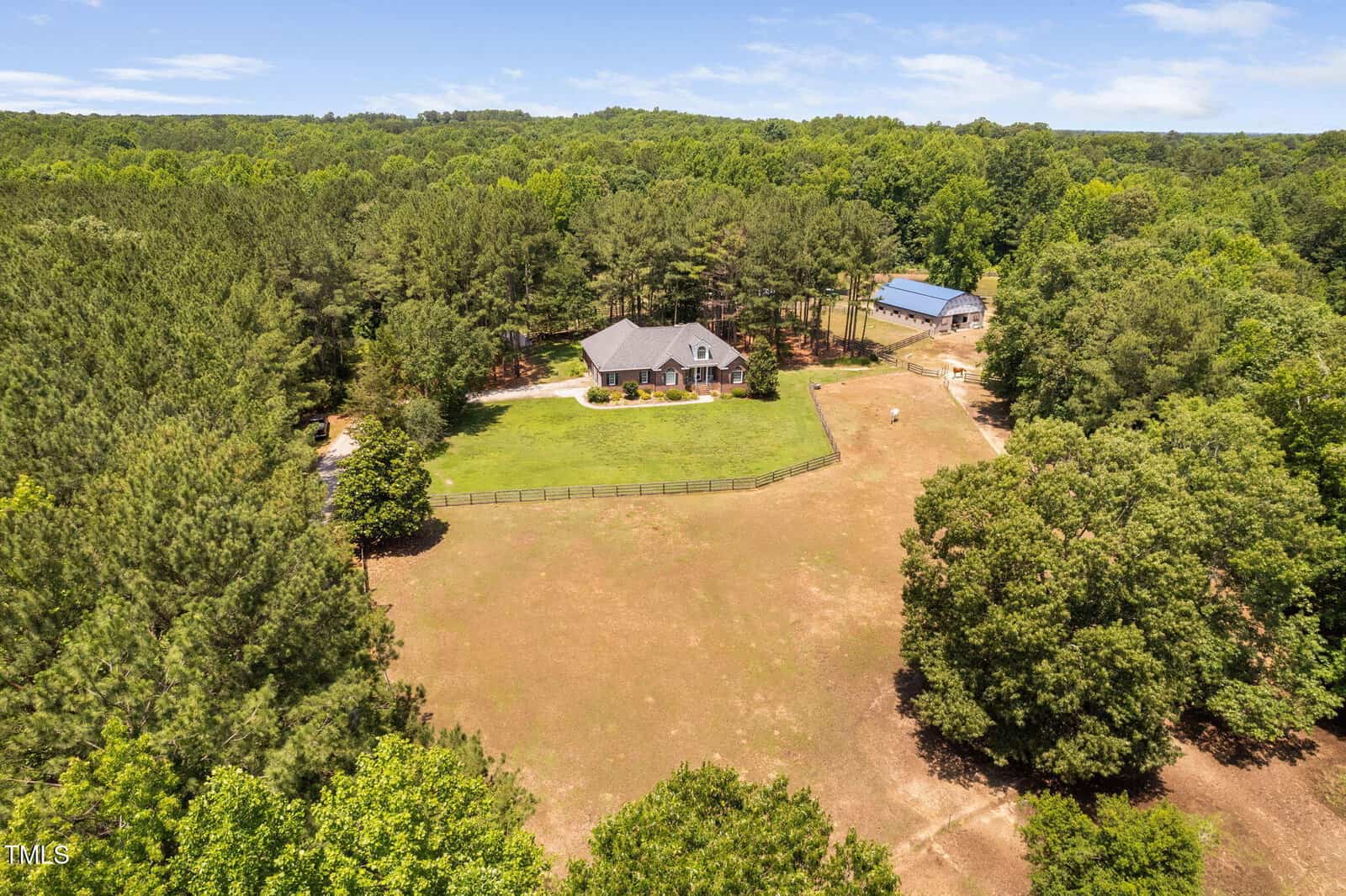Aerial view of a house surrounded by trees with a large grassy yard, fenced area, and a barn or outbuilding nearby, set in a rural, forested landscape under a partly cloudy sky.