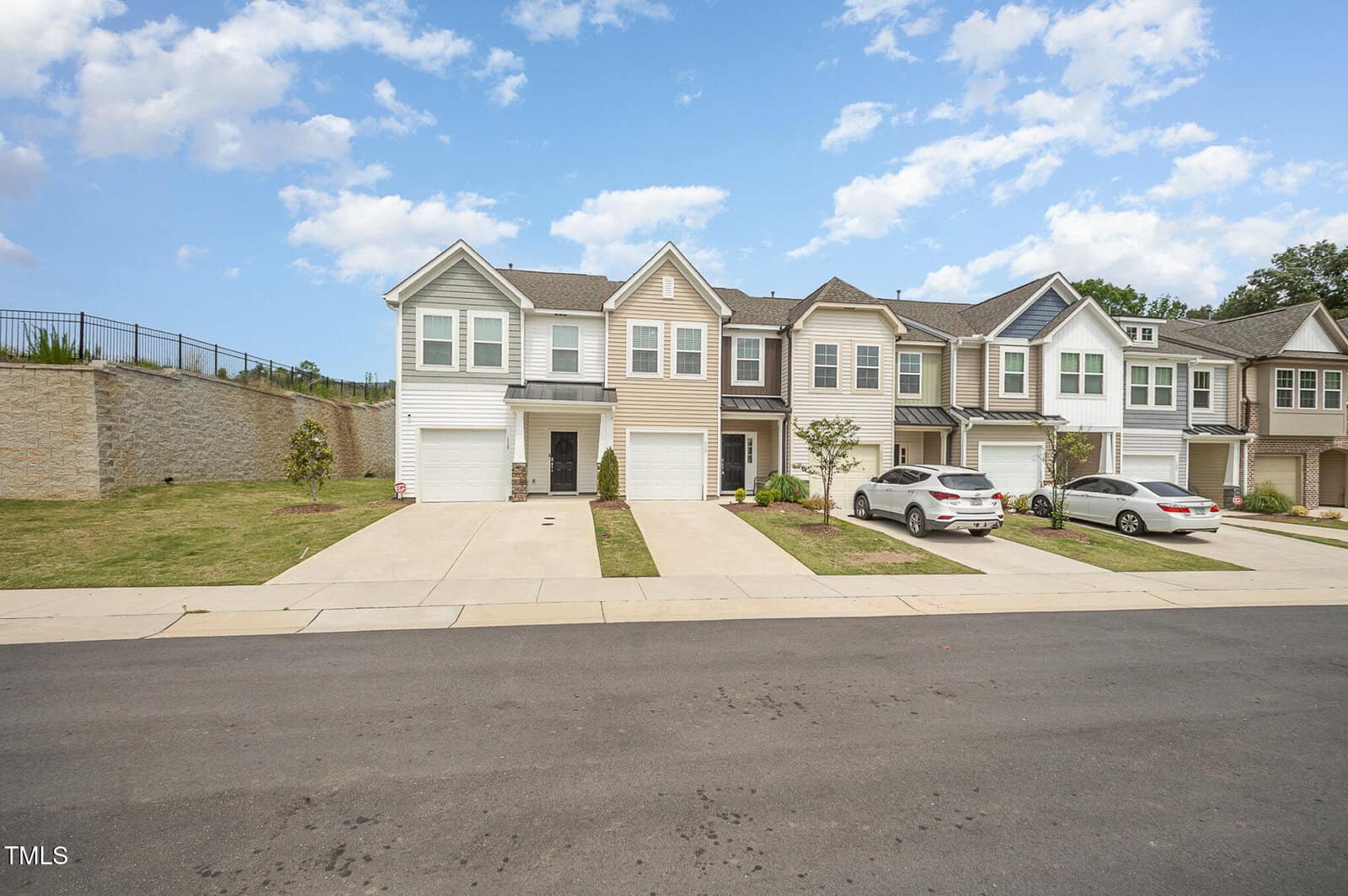 Row of modern two-story townhouses with attached garages and driveways, each with small lawns. Two cars are parked in front, and the sky is partly cloudy. A retaining wall and grassy area are on the left.