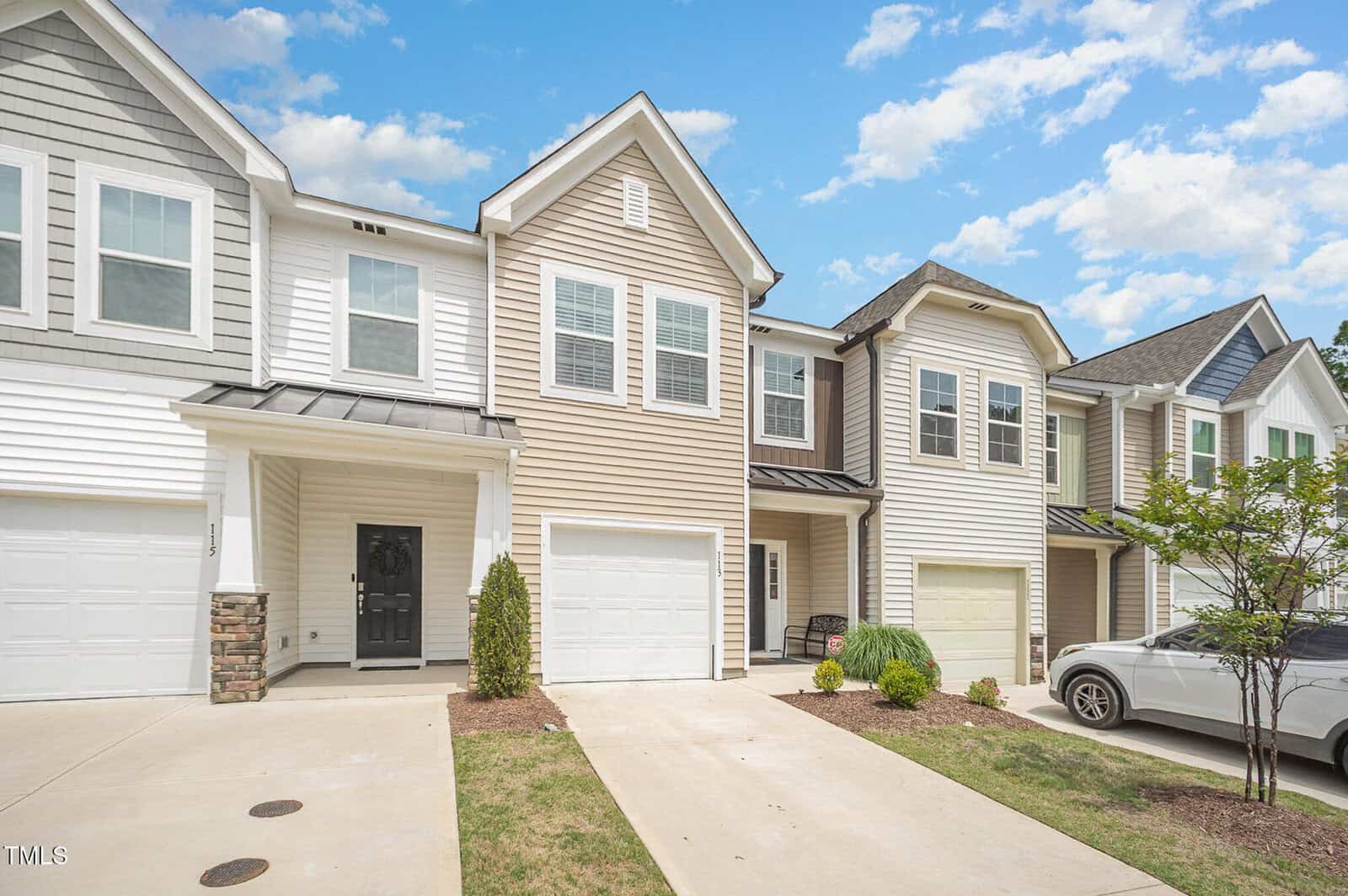 A row of modern two-story townhomes with beige and white siding, single-car garages, paved driveways, and small landscaped front yards under a blue sky with scattered clouds.