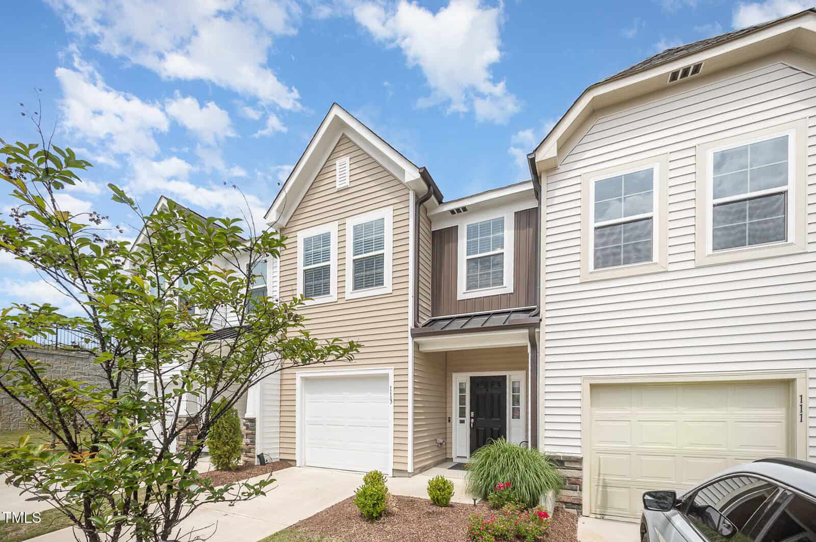 Two-story townhouse with beige and white siding, a single-car garage, and a black front door. Neatly landscaped front yard with bushes and a small tree under a partly cloudy sky.