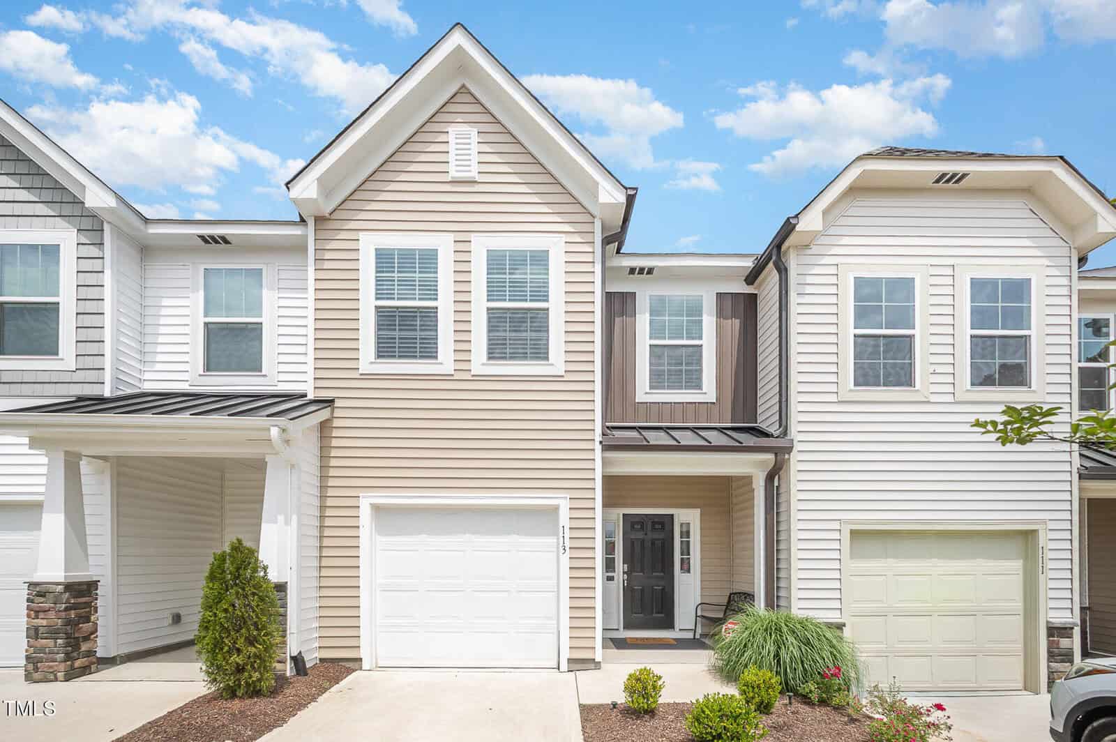 A modern two-story townhouse with tan and white siding, white trim, a single-car garage, and a black front door. The driveway and minimal landscaping are visible under a blue sky with scattered clouds.