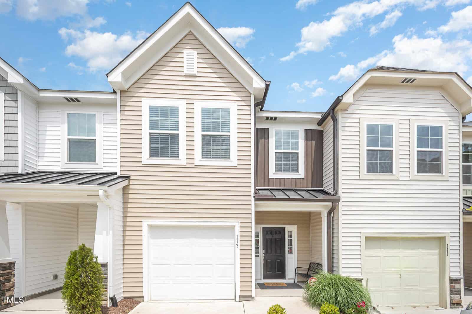 A two-story townhouse with beige and white siding, black door, and single-car garage. There are two large windows above the garage and entrance, and small landscaped bushes by the driveway. The sky is bright with scattered clouds.