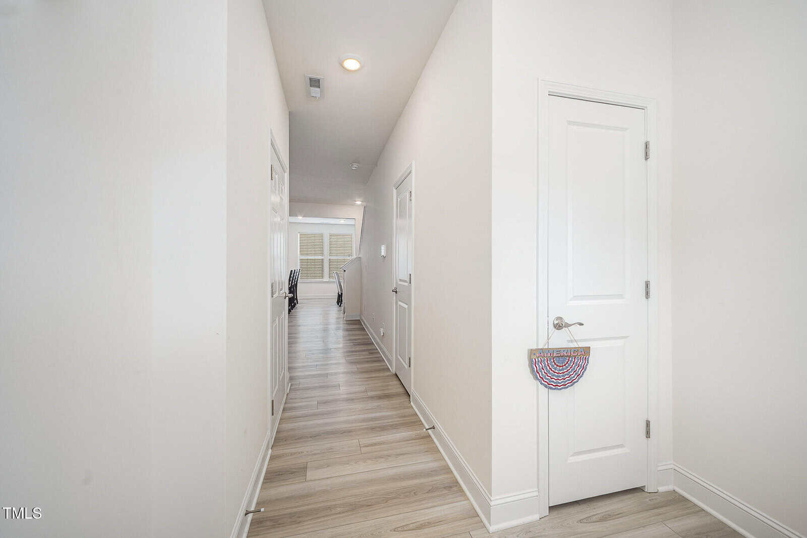 A bright hallway with light wood floors, white walls, and several doors. One door on the right has a child’s rainbow basket hanging from the handle. The hallway leads to a living area with windows.