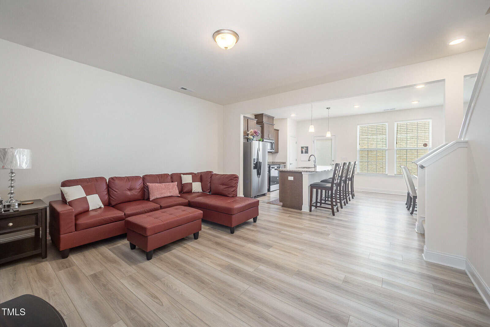 Open-concept living area with a red sectional sofa, side table, and lamp. Kitchen with island, stainless steel appliances, and bar stools in the background. Dining area with table and chairs near two large windows.