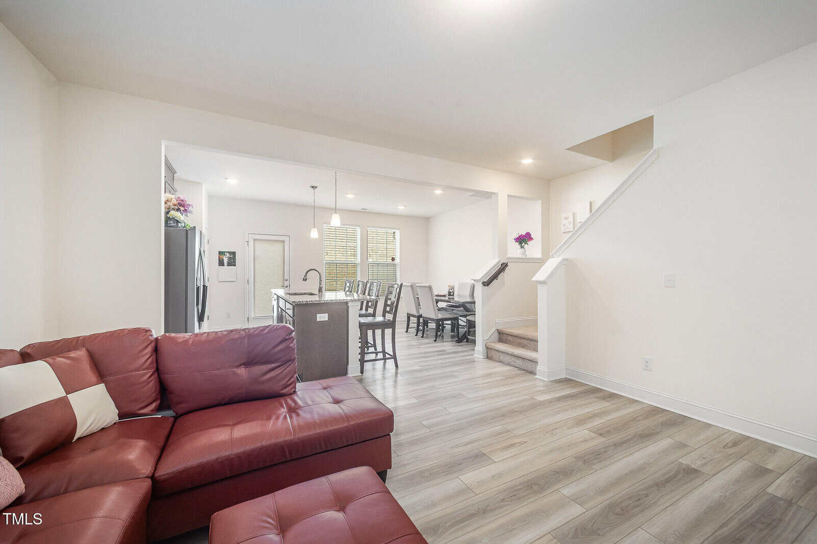 Open floor plan with a red sectional sofa, wood-style flooring, a modern kitchen with an island, dining table, and staircase leading up, in a well-lit living space.