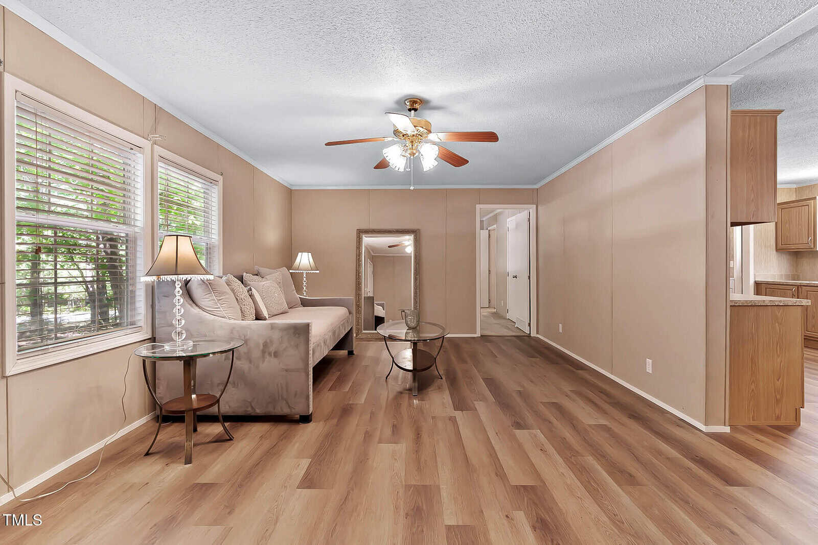 A living room with wood flooring, beige walls, a ceiling fan with lights, a beige sofa, glass side and coffee tables, a lamp, a wall mirror, and windows with blinds. The kitchen is visible through an open doorway.