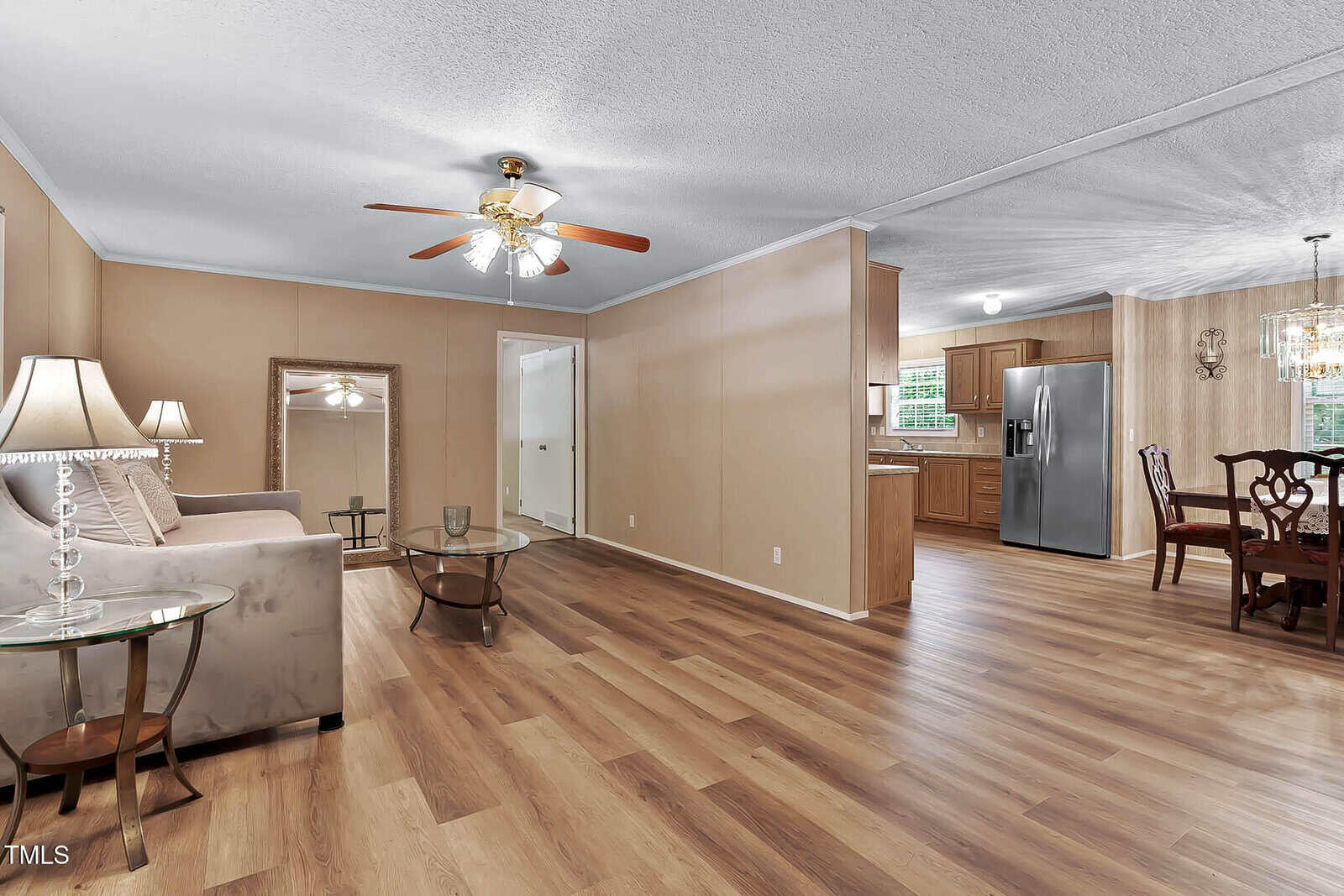 Living room with wood flooring, beige walls, a ceiling fan, sofa, glass tables, and a large mirror. Dining area and kitchen with stainless steel refrigerator are visible in the background.