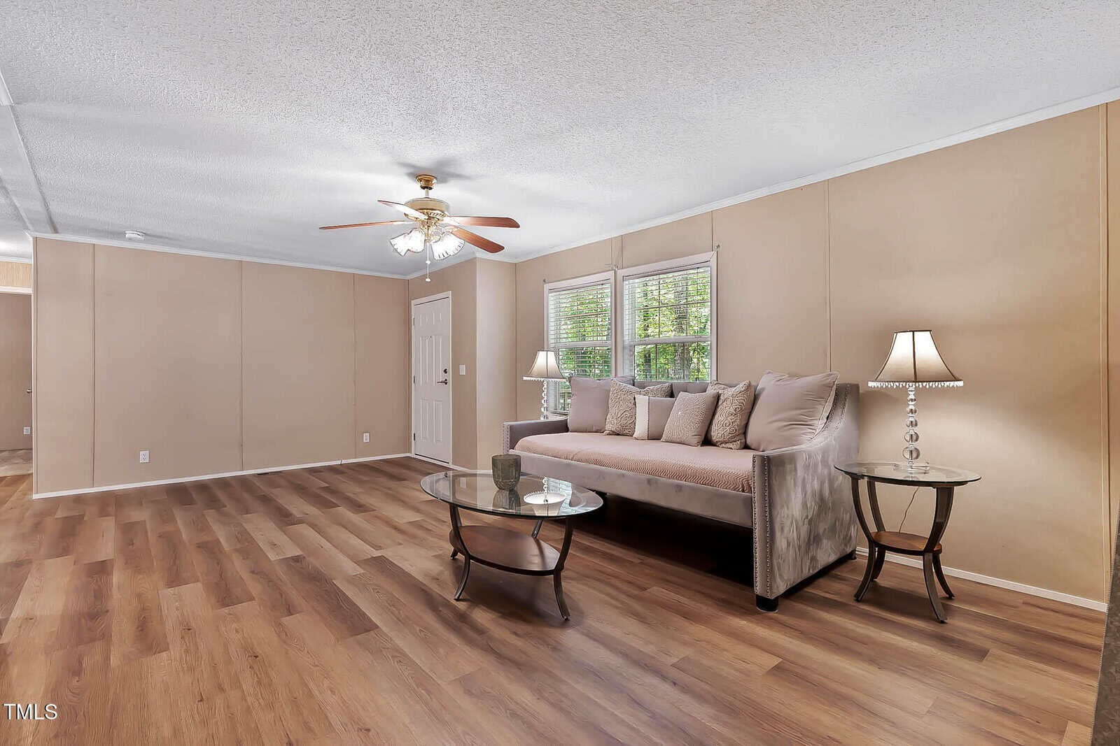 A spacious living room with wood flooring, beige walls, a gray sofa with multiple pillows, two glass side tables with lamps, a glass coffee table, and a ceiling fan with lights. Two windows let in natural light.