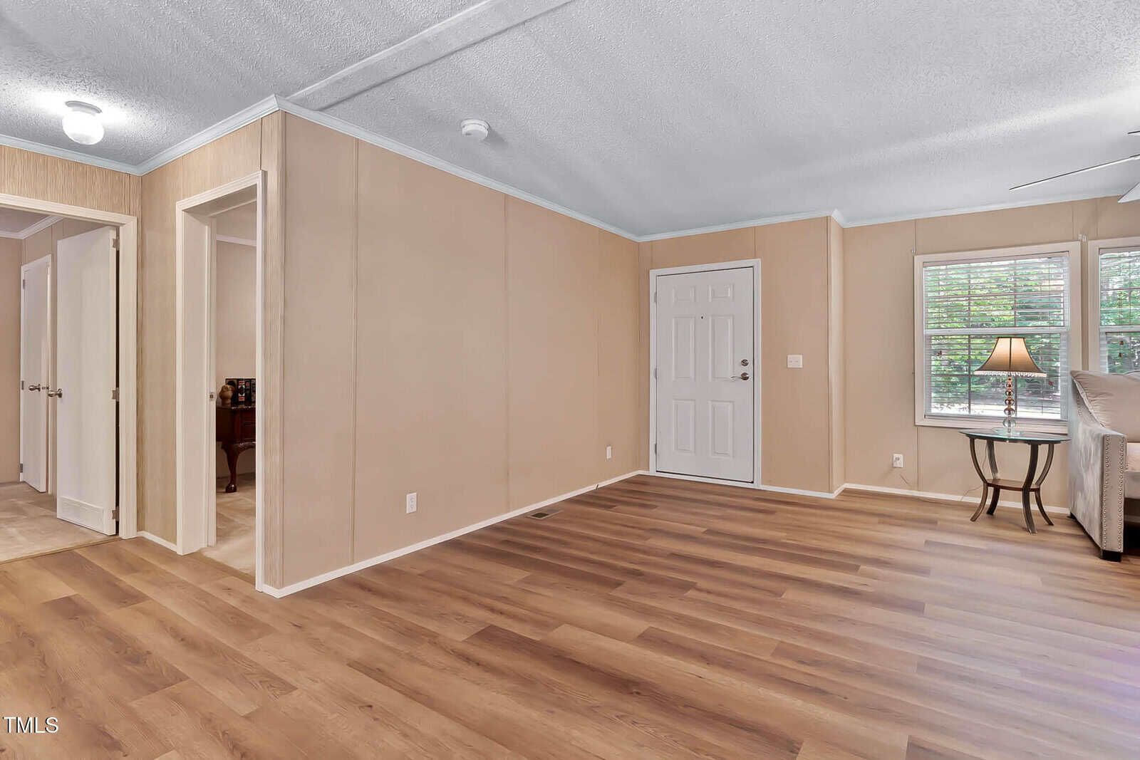 A room with light wood flooring, beige walls, a white door, and two open doorways. To the right, a window with blinds and a side table with a lamp can be seen next to a sofa. Ceiling has a beam and visible light fixture.
