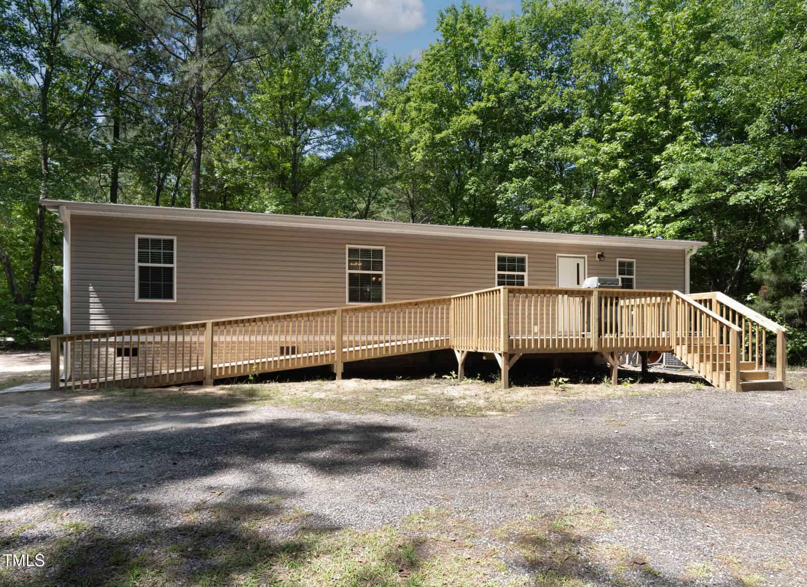 A beige single-story modular home with white trim, surrounded by trees. A wooden accessibility ramp leads to the front door. The sky is blue with some clouds, and the ground is covered with gravel and grass.