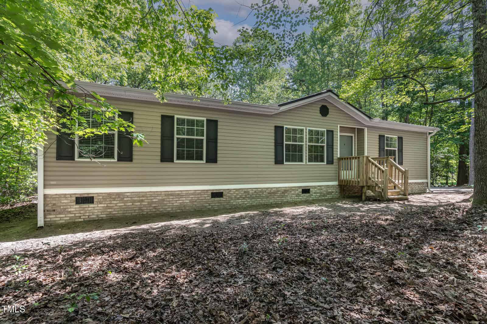A beige, single-story house with black shutters and a small wooden porch is surrounded by dense green trees and leafy ground cover on a sunny day.