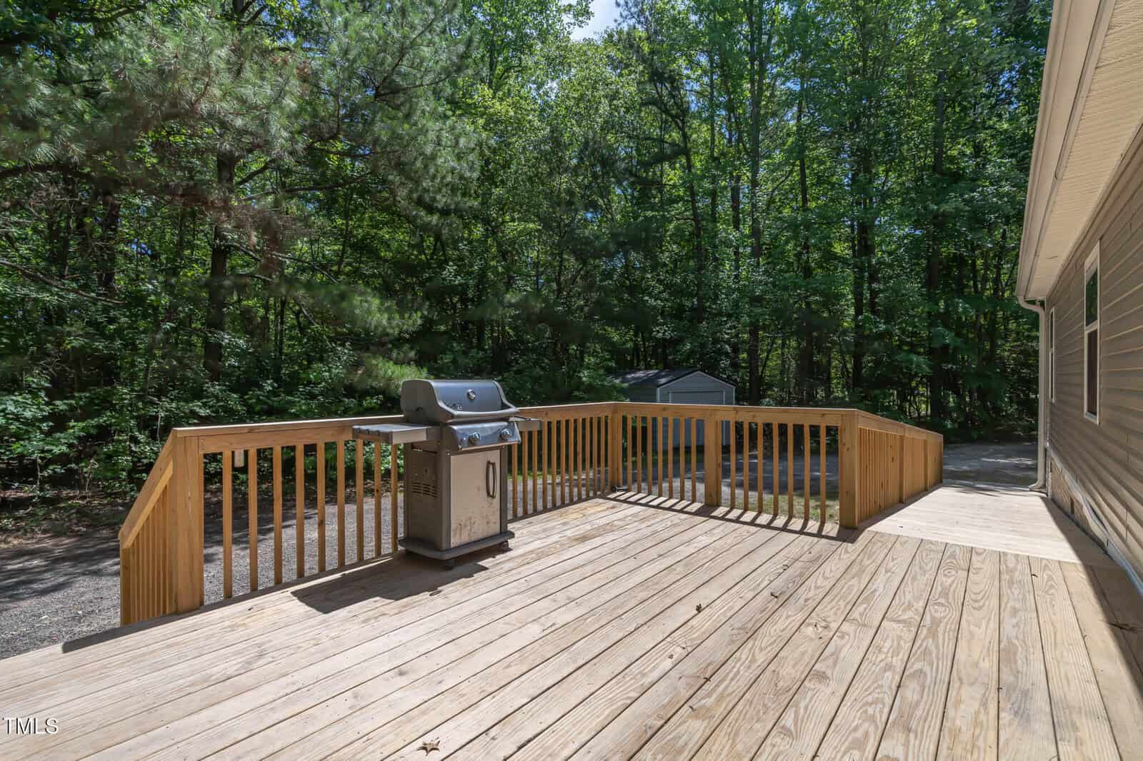A wooden deck with a gas grill overlooks a wooded backyard with tall trees and a small gray shed in the background on a sunny day.