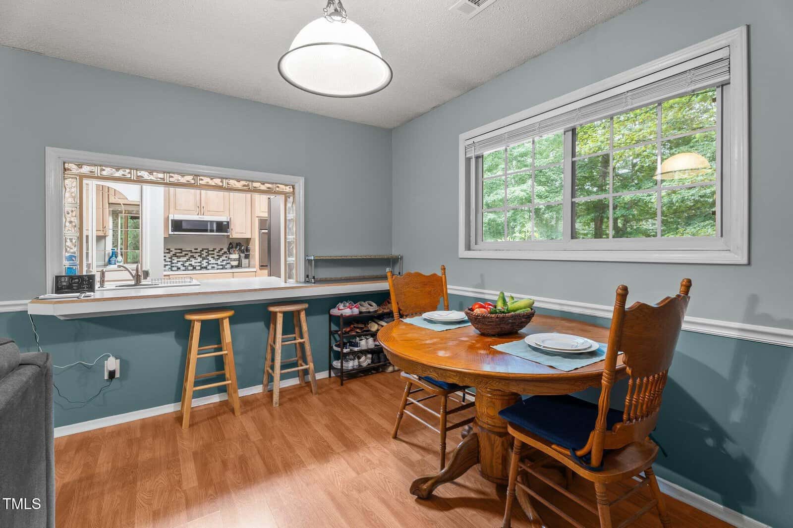 A small dining area with a round wooden table set for two, two wooden chairs, wood flooring, a window showing greenery, and a pass-through to a kitchen with stools on the other side.