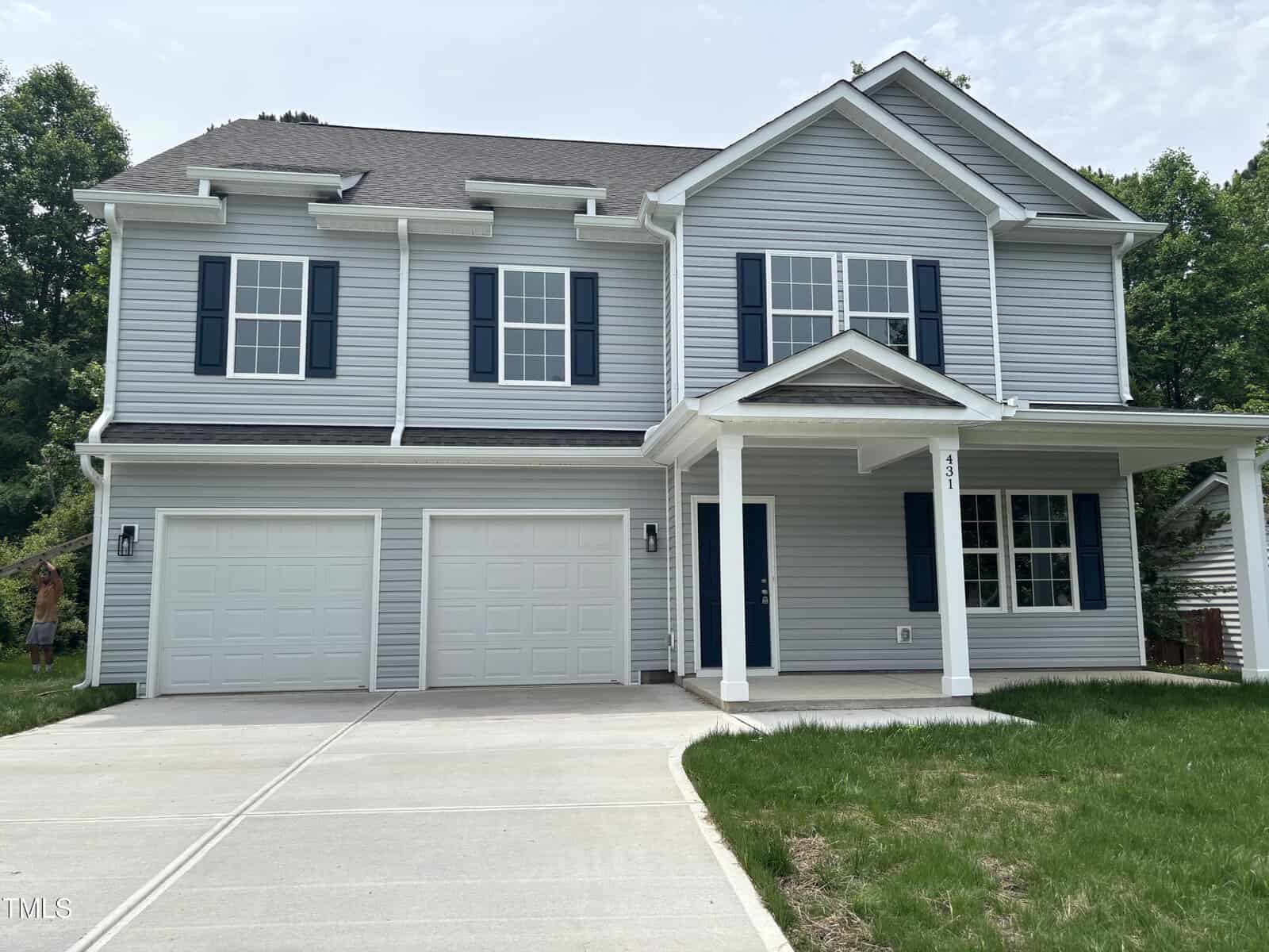 A two-story gray house with blue shutters and a covered front porch. There are two garage doors, a large driveway, and a small lawn area with minimal landscaping. Trees are visible in the background.