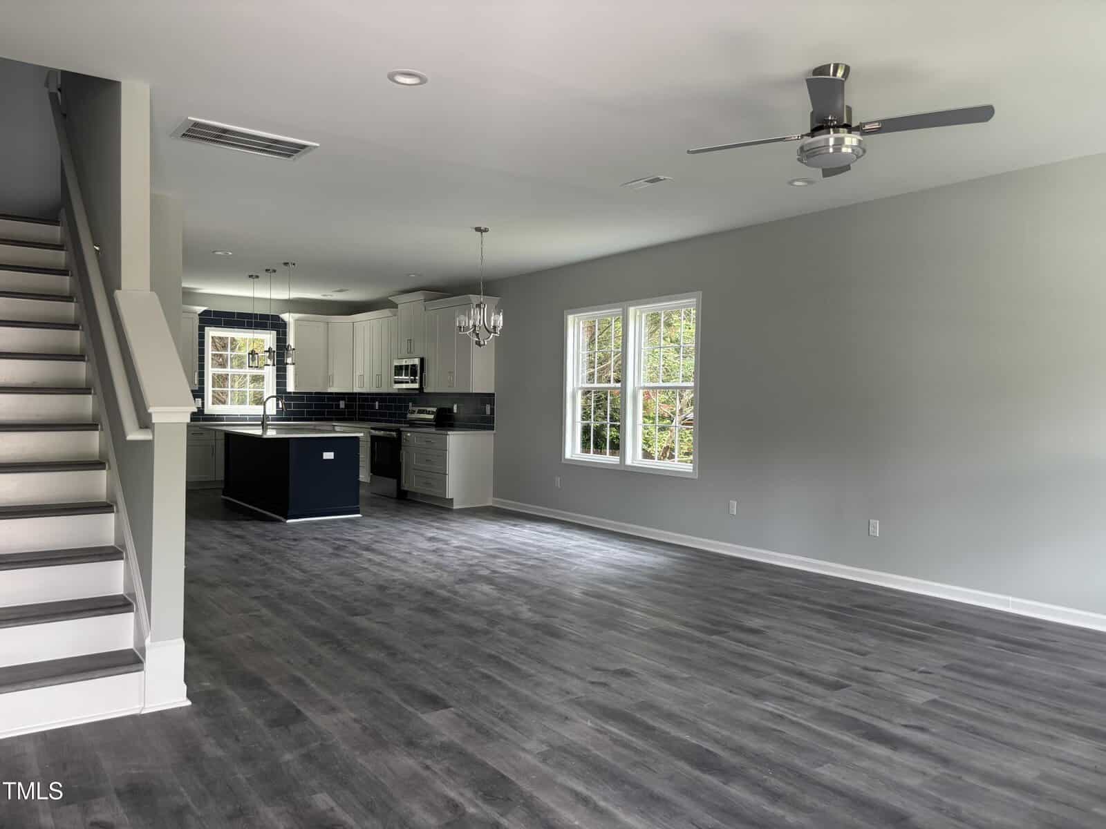 Open-concept living area with gray flooring, light gray walls, large windows, a staircase on the left, and a modern kitchen with white cabinets, a dark island, and stainless steel appliances in the background.