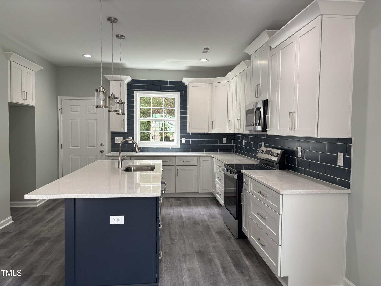 Modern kitchen with white cabinets, navy blue subway tile backsplash, stainless steel appliances, a large island with a sink, pendant lighting, and gray wood-look flooring. A window is centered above the sink.