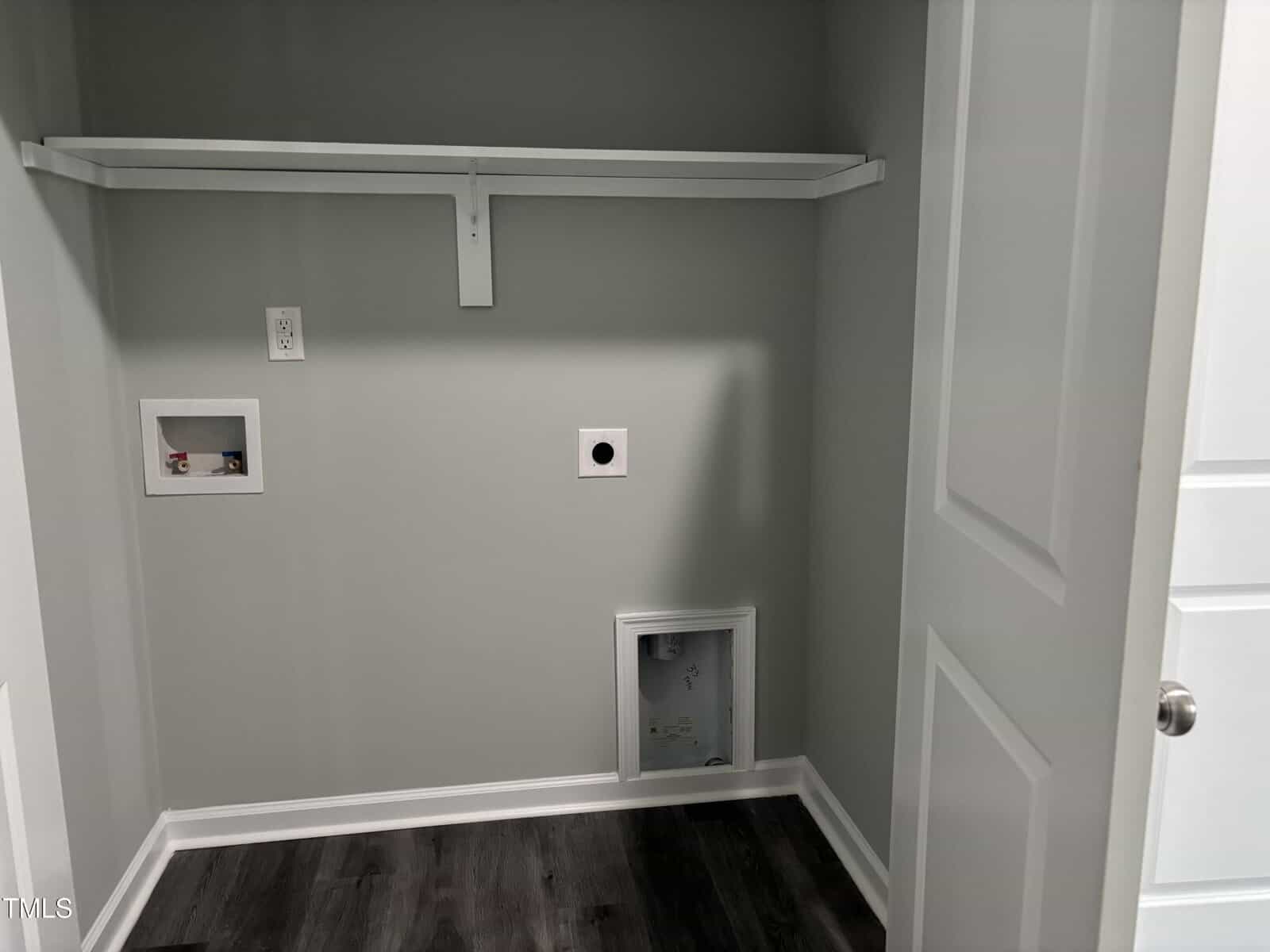 A laundry room with light gray walls, white trim, dark wood flooring, plumbing hookups for a washer and dryer, an electrical outlet, and a white shelf mounted on the wall.