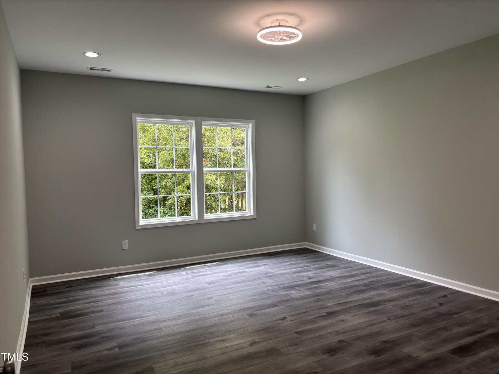 Empty room with light gray walls, a ceiling light, two windows with white trim, and dark wood-style flooring. Room appears clean and well-lit with natural light coming in from the windows.