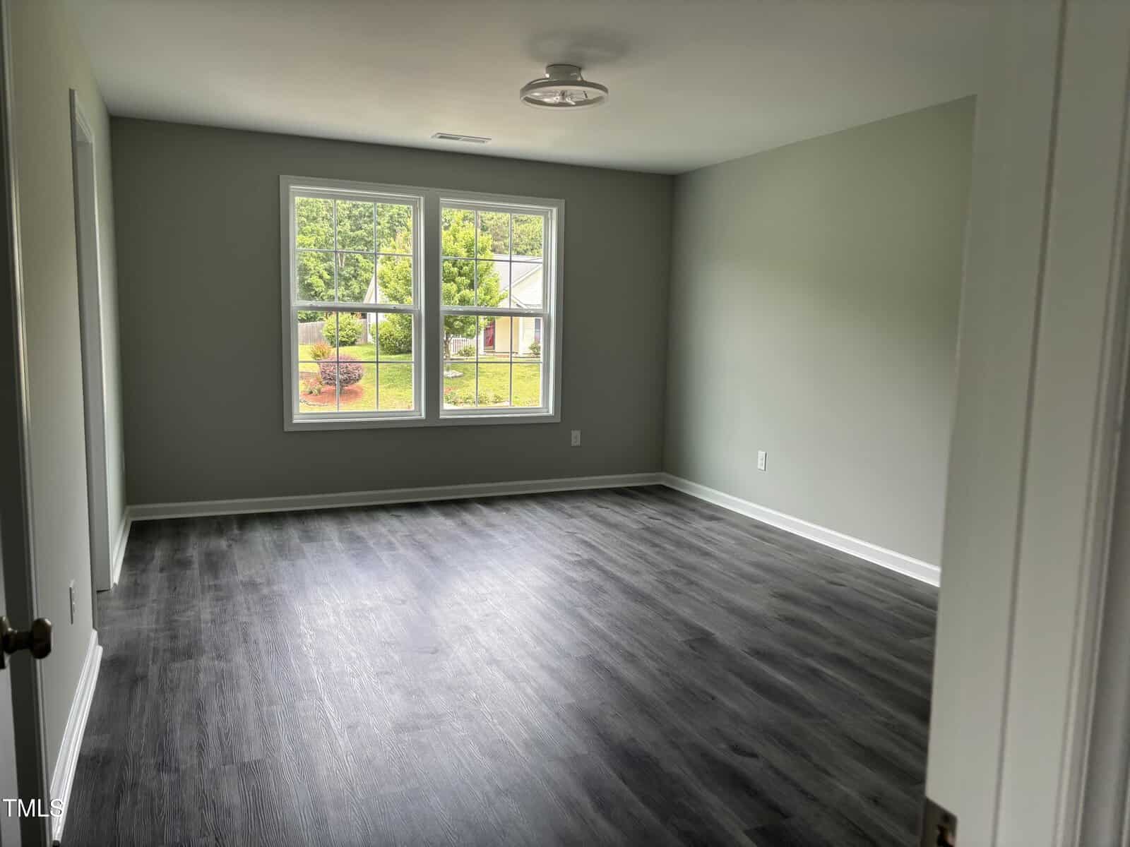 An empty room with gray walls, dark wood-style flooring, a ceiling light fixture, and a large window showing a view of trees and a house outside. The room appears clean and well-lit.