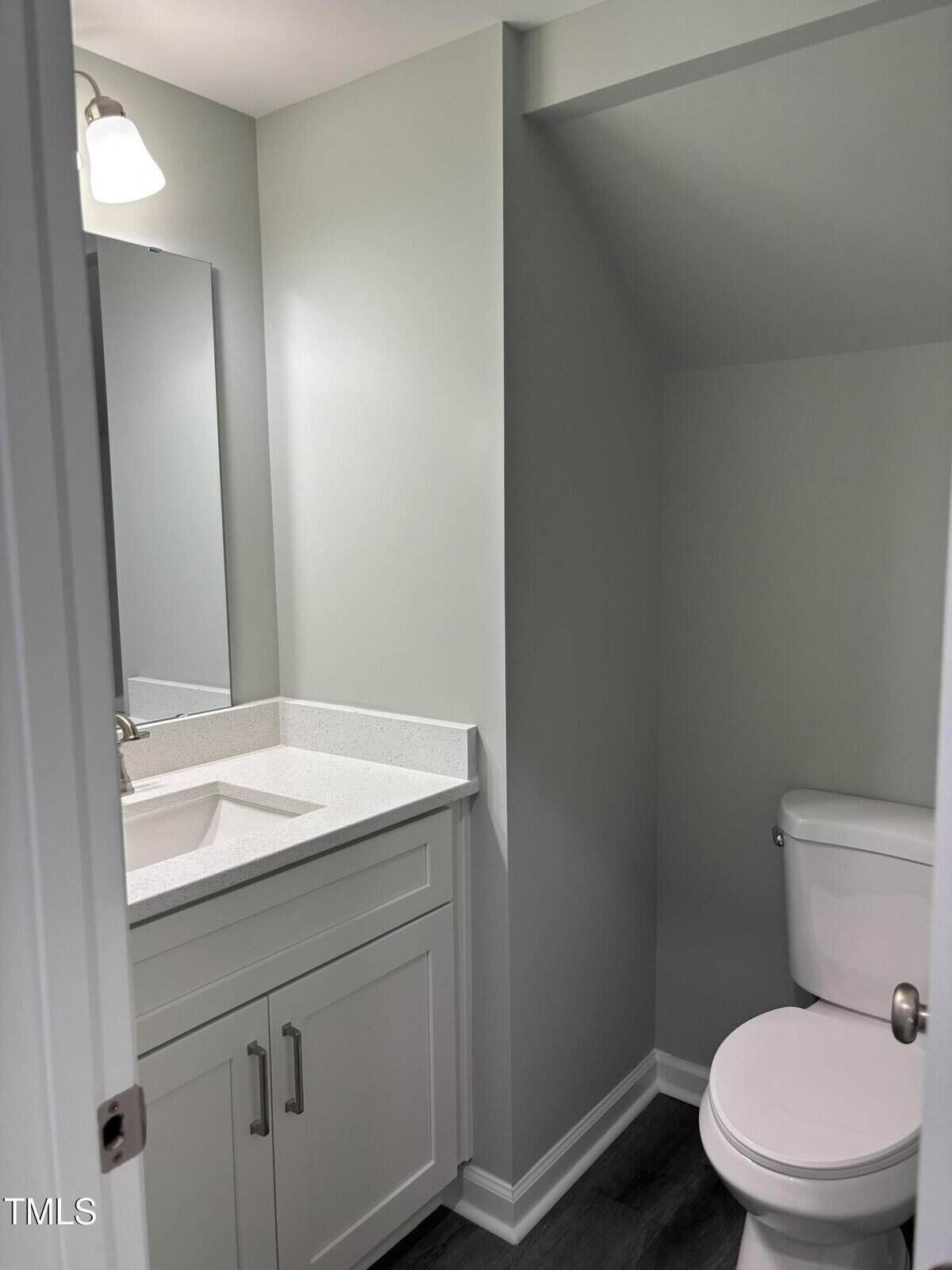 A small bathroom with light gray walls, a white vanity with a sink and countertop, a rectangular mirror above, and a toilet positioned under a sloped ceiling. The floor is dark wood or laminate.