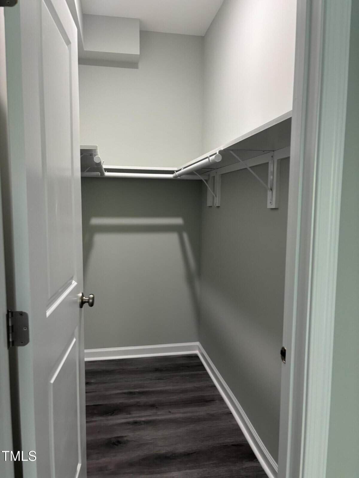 A small walk-in closet with light gray walls, white trim, and dark wood flooring. White shelving and hanging rods are installed on the left and back walls. The door is partially open.