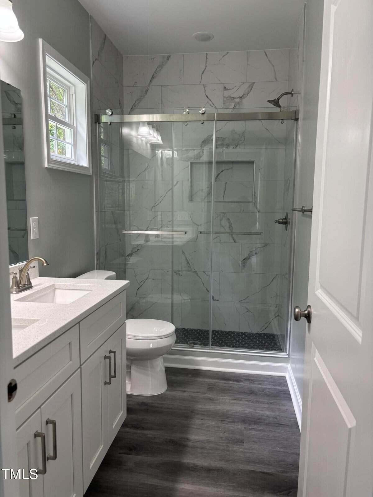 A modern bathroom with white cabinets, a white countertop with dual sinks, a toilet, and a glass-enclosed shower with marble-patterned walls and dark tile flooring. A small window lets in natural light.