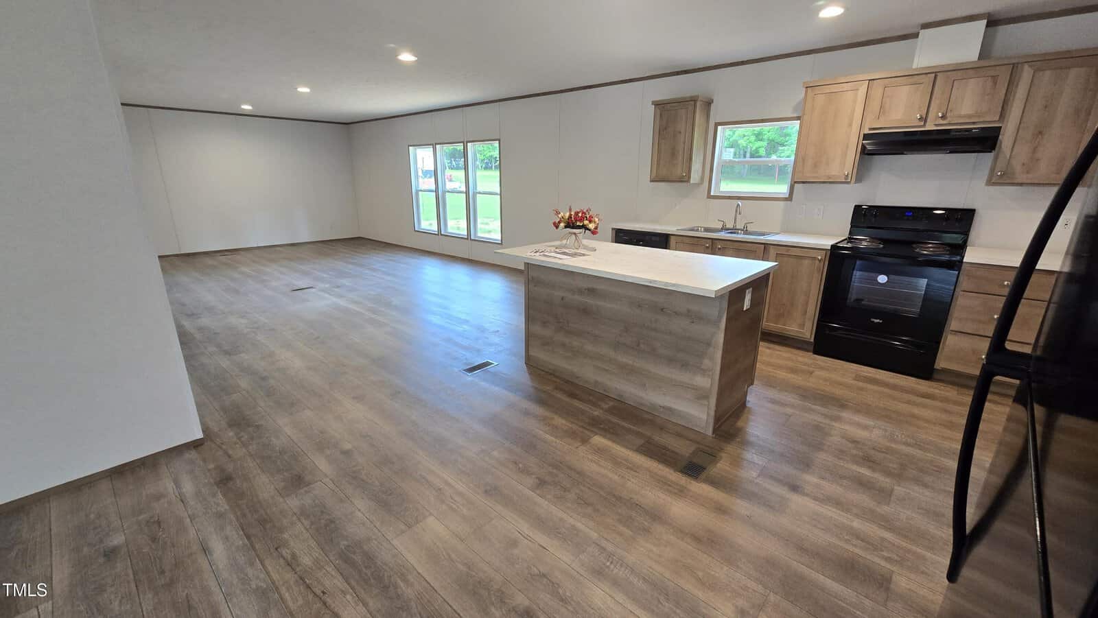 Spacious open-plan kitchen and living area with wood-look flooring, light wood cabinets, black appliances, white countertops, and large windows letting in natural light. Room appears unfurnished except for a dish arrangement on the island.