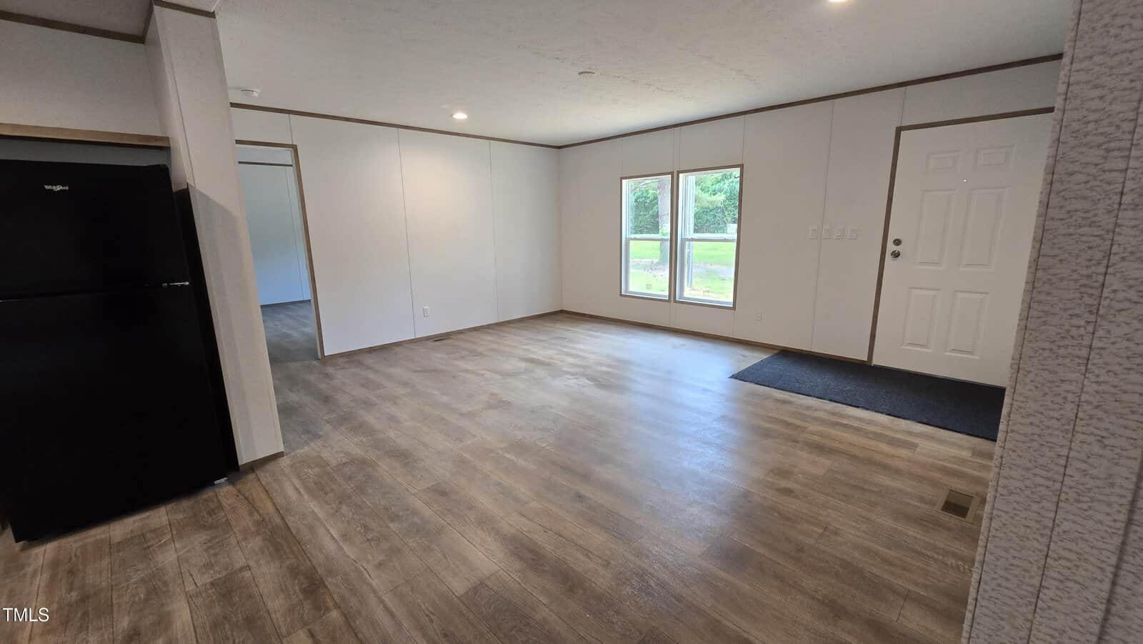 An empty room with wood-style flooring, white walls, a white door, two windows, and a black refrigerator partially visible on the left. Natural light enters through the windows, and greenery is visible outside.