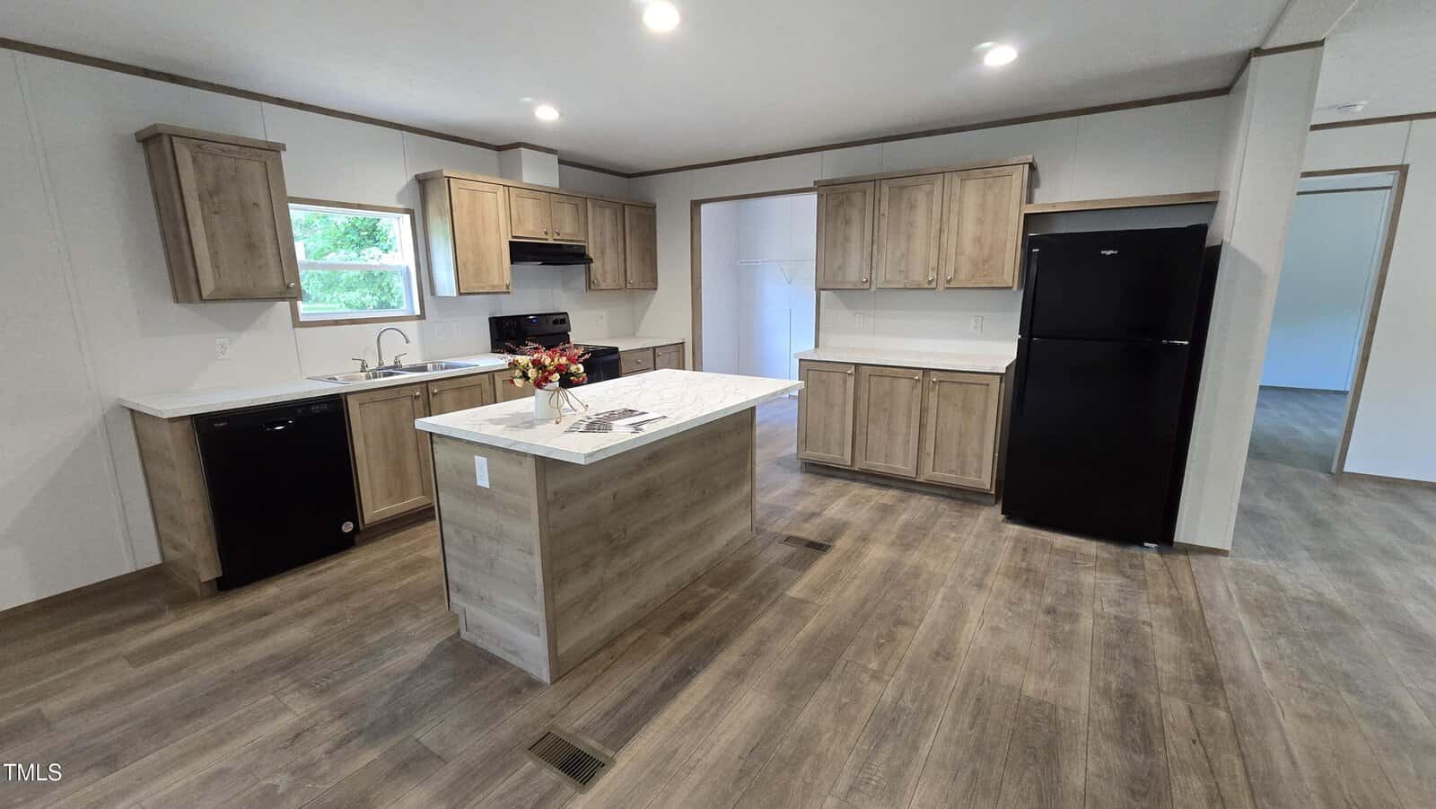 A modern kitchen with wood cabinets, black appliances including a refrigerator, stove, and dishwasher, a center island with marble-style countertop, and wood-look flooring. A vase with flowers sits on the island.