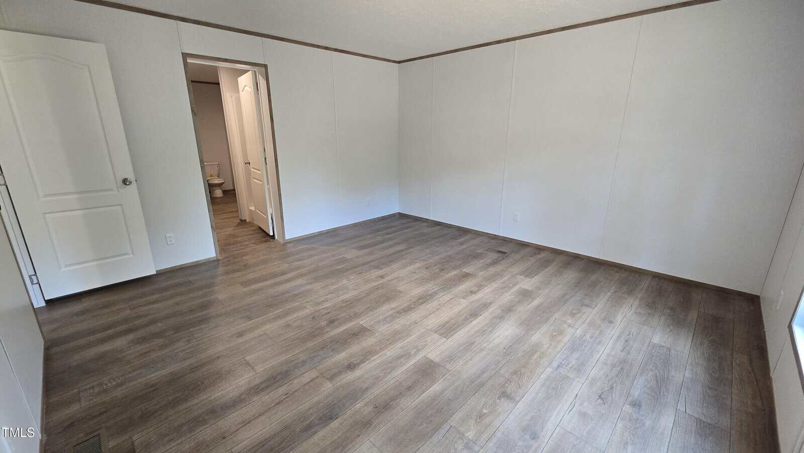 An empty room with light-colored walls, wood-look flooring, a window on the right, and two doors on the left wall, one slightly open revealing a bathroom.