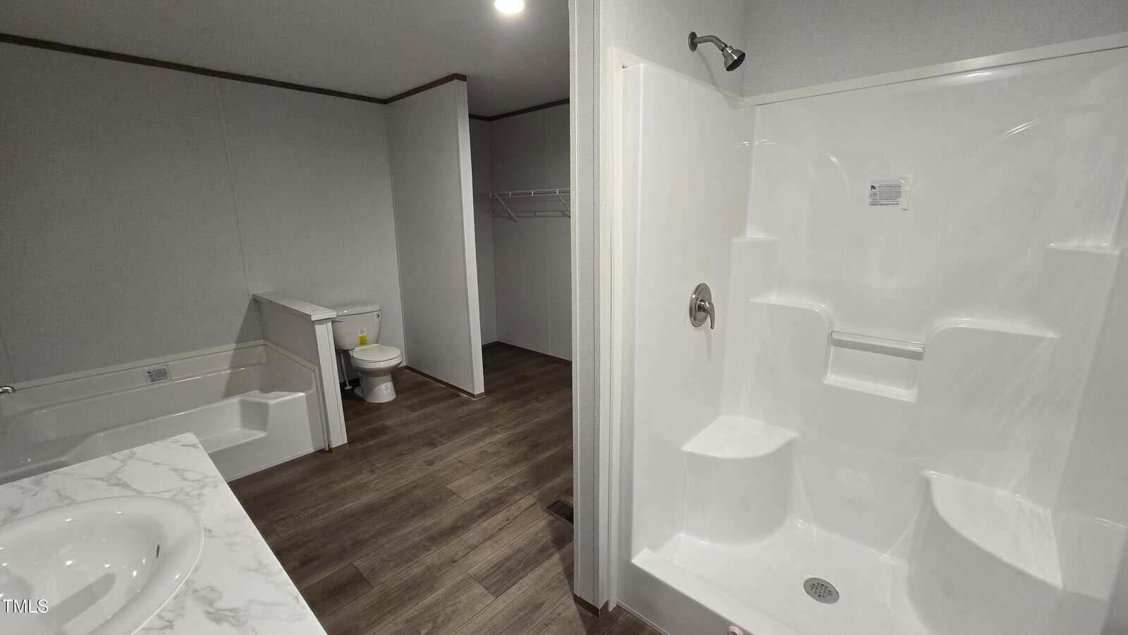 A bathroom with a white bathtub, a toilet, a walk-in shower with built-in seats, a vanity with a marble countertop, wood-style flooring, and an open closet area in the background.