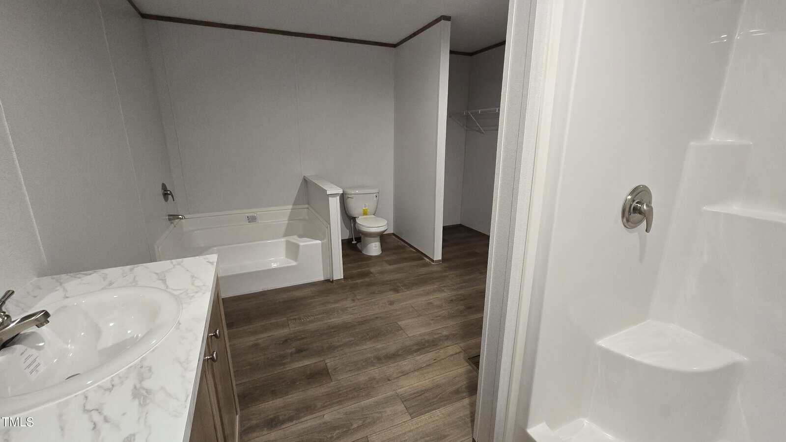 A bathroom with a marble countertop sink, a bathtub, a toilet, a separate shower, and a walk-in closet area; the floor is wood-patterned, and the walls are plain white.