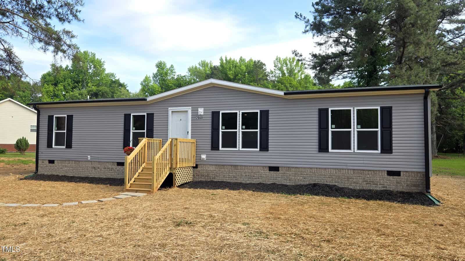 A single-story gray manufactured home with black shutters, white trim, and a small wooden staircase leading to the front door, set on a lot with straw-covered ground and bordered by trees.
