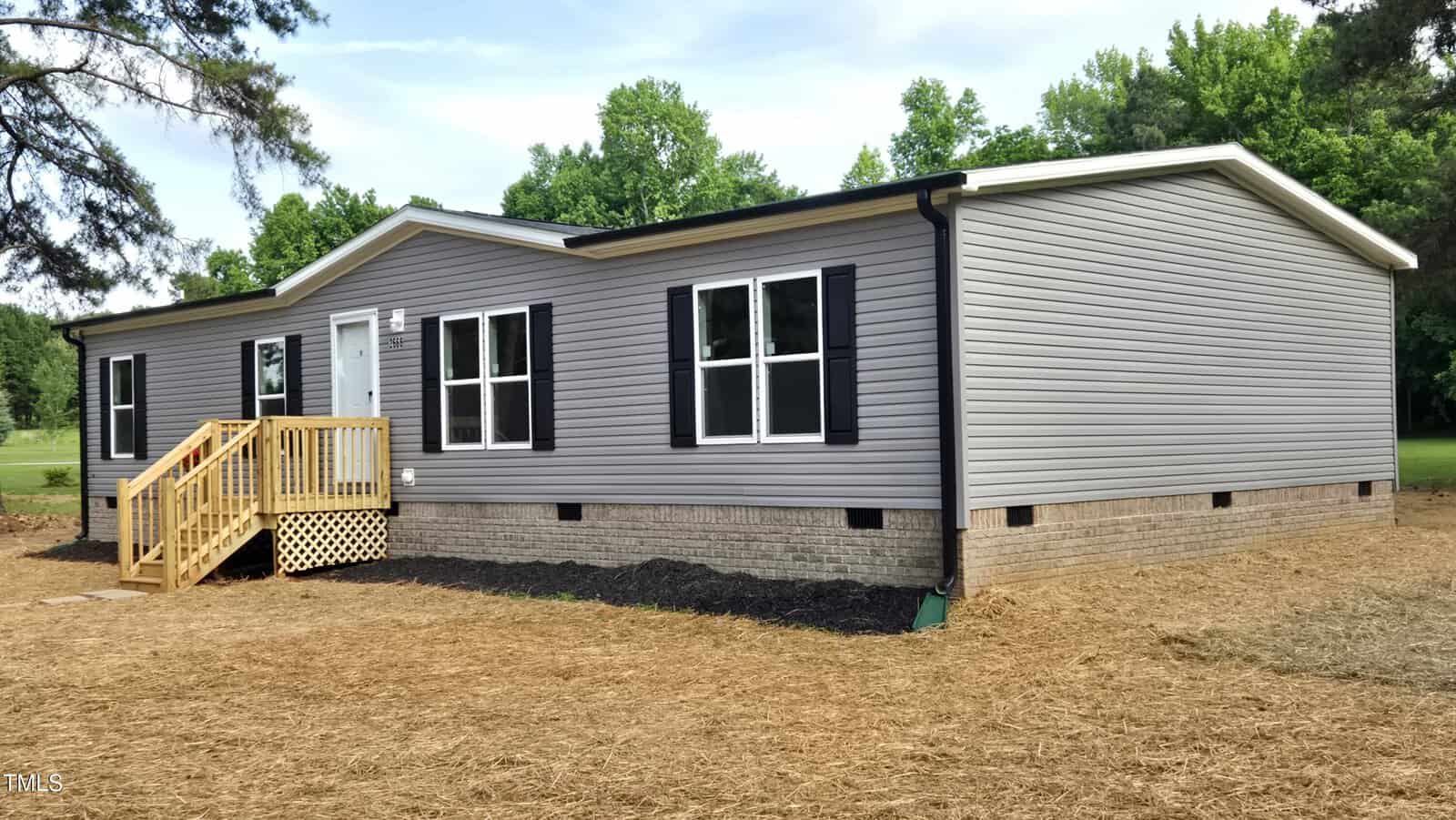 A single-story gray manufactured home with black shutters and a small wooden front porch sits on a lot with straw-covered ground and trees in the background.