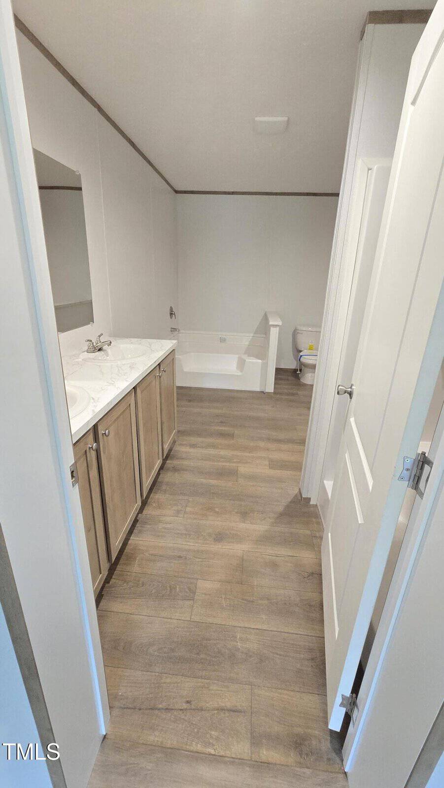 A bathroom with wood-look flooring, a double sink vanity with a marble countertop and mirror on the left, a bathtub against the back wall, and a toilet next to the tub. The room has neutral wall colors.