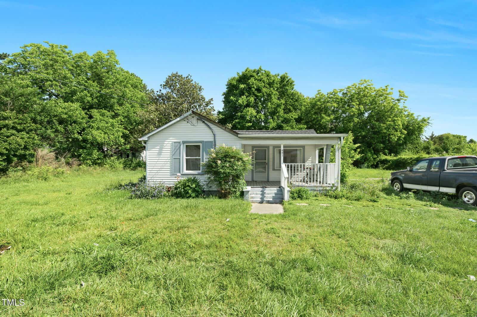 A small white single-story house at 441 Cedar Street, Henderson, features a covered front porch and sits on a large grassy lot surrounded by trees. A black pickup truck is parked to the right under a clear blue sky.