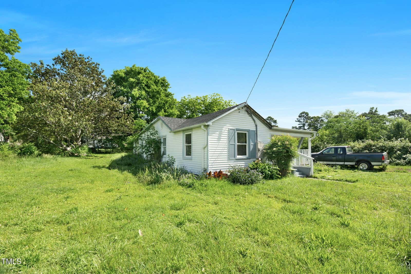 A small white house with a front porch sits on a grassy lawn with trees in the background. A black pickup truck is parked nearby under a clear blue sky.