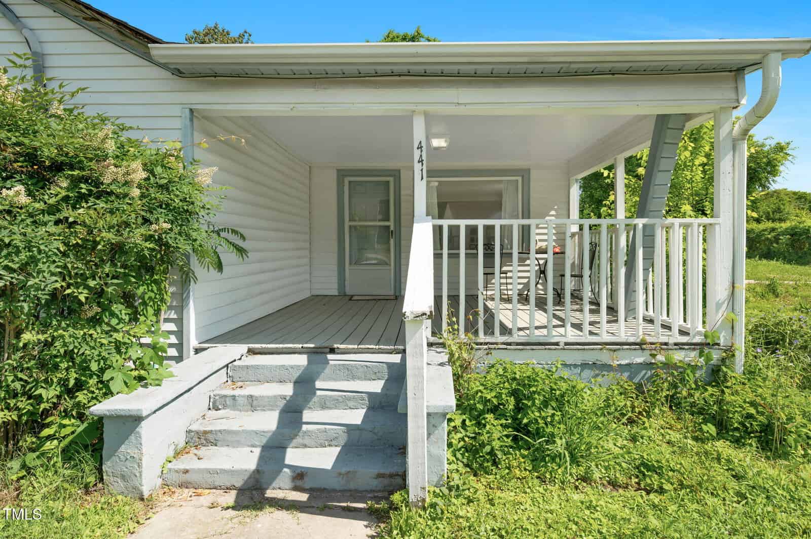 Front view of a white house with a covered porch, steps leading up, a railing, and overgrown greenery on either side. The house has two visible doors and a light fixture by the entrance.