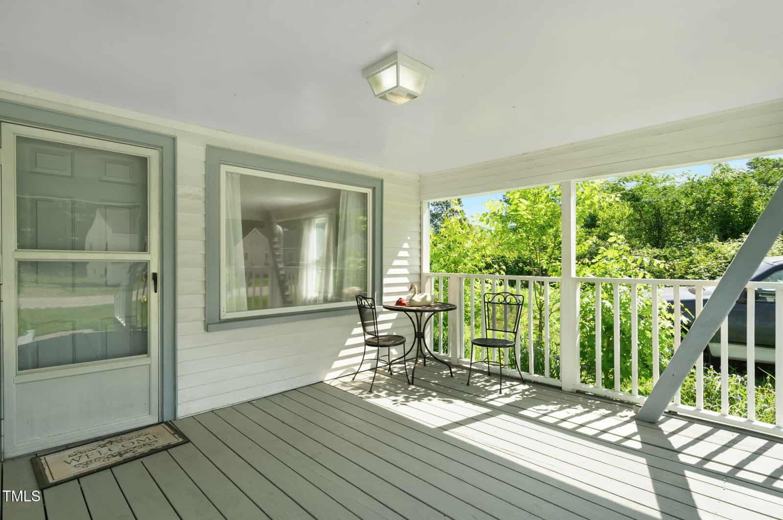 A covered porch with gray flooring, white railings, a gray-trimmed door, a large window, and a small black metal table with two matching chairs. Sunlight filters in and greenery is visible beyond the railing.
