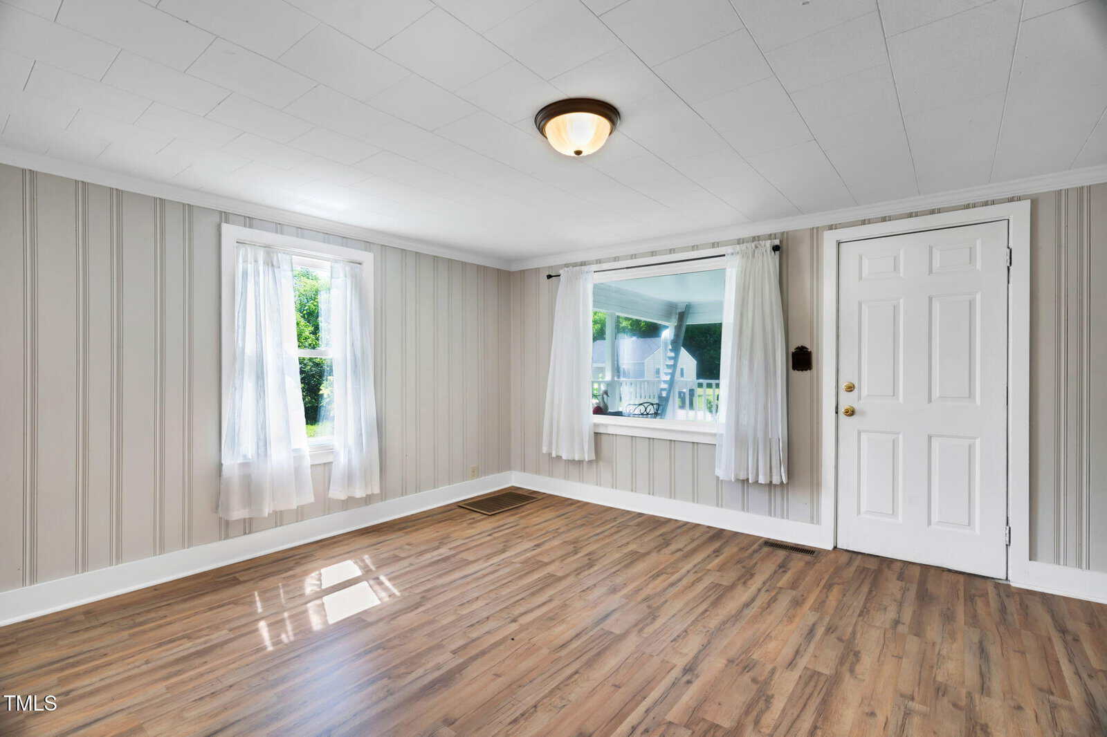 Empty room with light-colored wood flooring, two windows with sheer white curtains, beige vertical wall paneling, a white ceiling with a single light fixture, and a white front door.