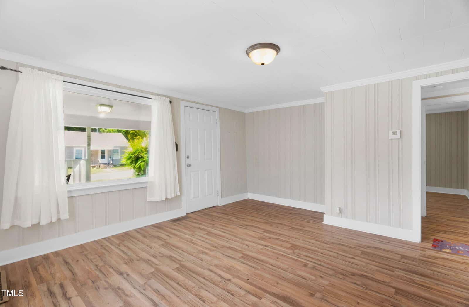 A small, empty room with light-colored wood flooring, beige striped walls, a white ceiling, a single ceiling light, a window with white curtains, and a white door leading outside.