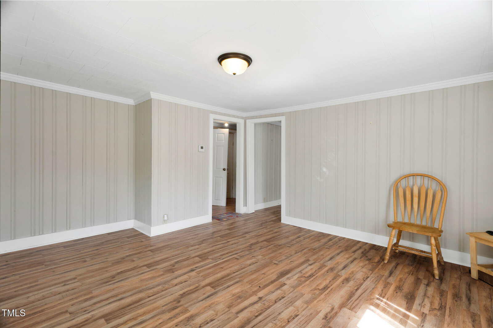 A mostly empty room with light wood flooring, gray vertical panel walls, white baseboards and trim, a ceiling light, a single wooden chair, and a small table near a doorway leading to another room.