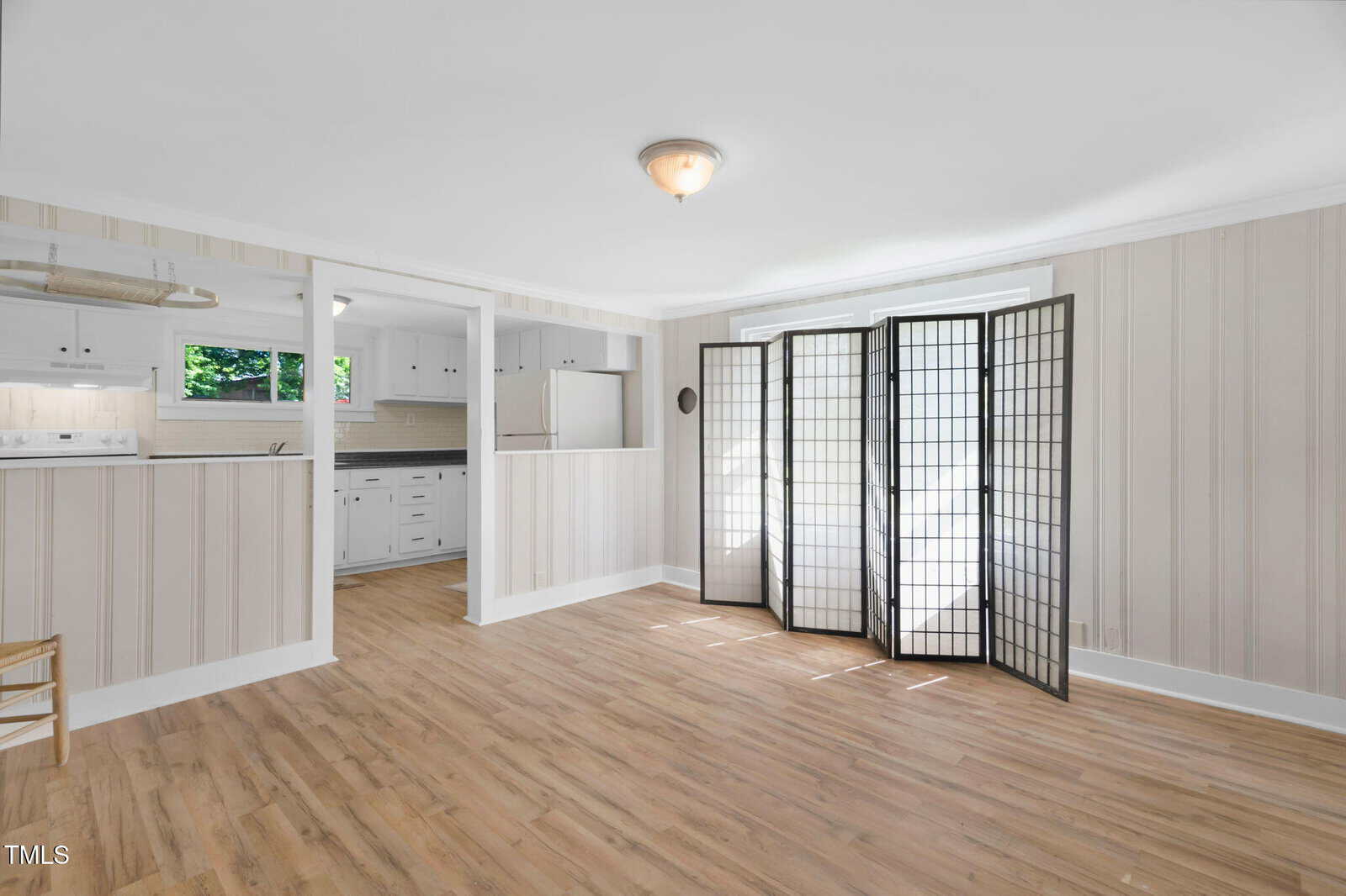 A bright room with light wood flooring, striped wallpaper, a ceiling light, a folding room divider with white panels, and an open view into a white kitchen with cabinetry and a window.