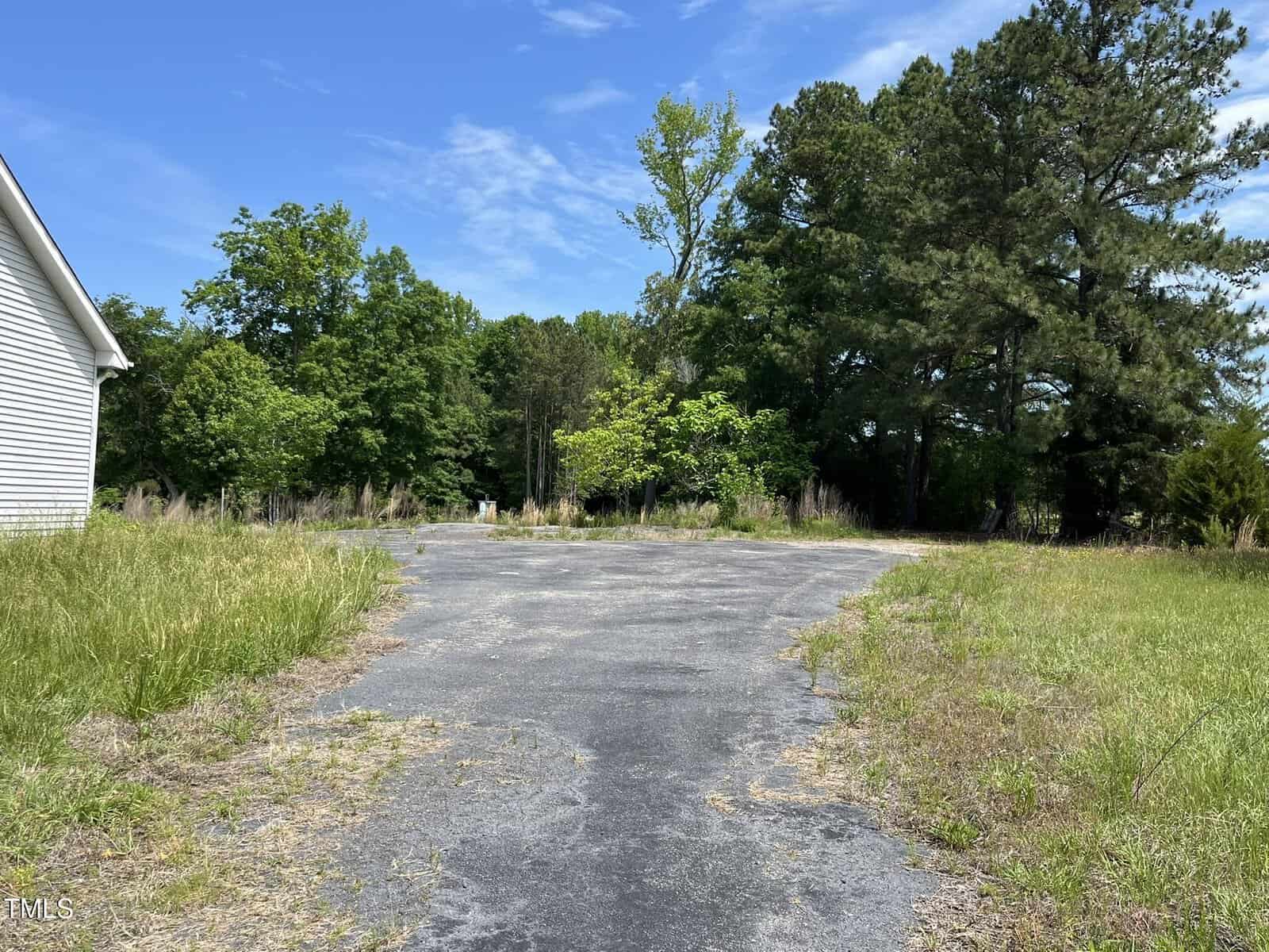 A paved driveway with cracks and overgrown grass on both sides leads toward a wooded area with tall green trees under a partly cloudy blue sky. The side of a white building is visible on the left.