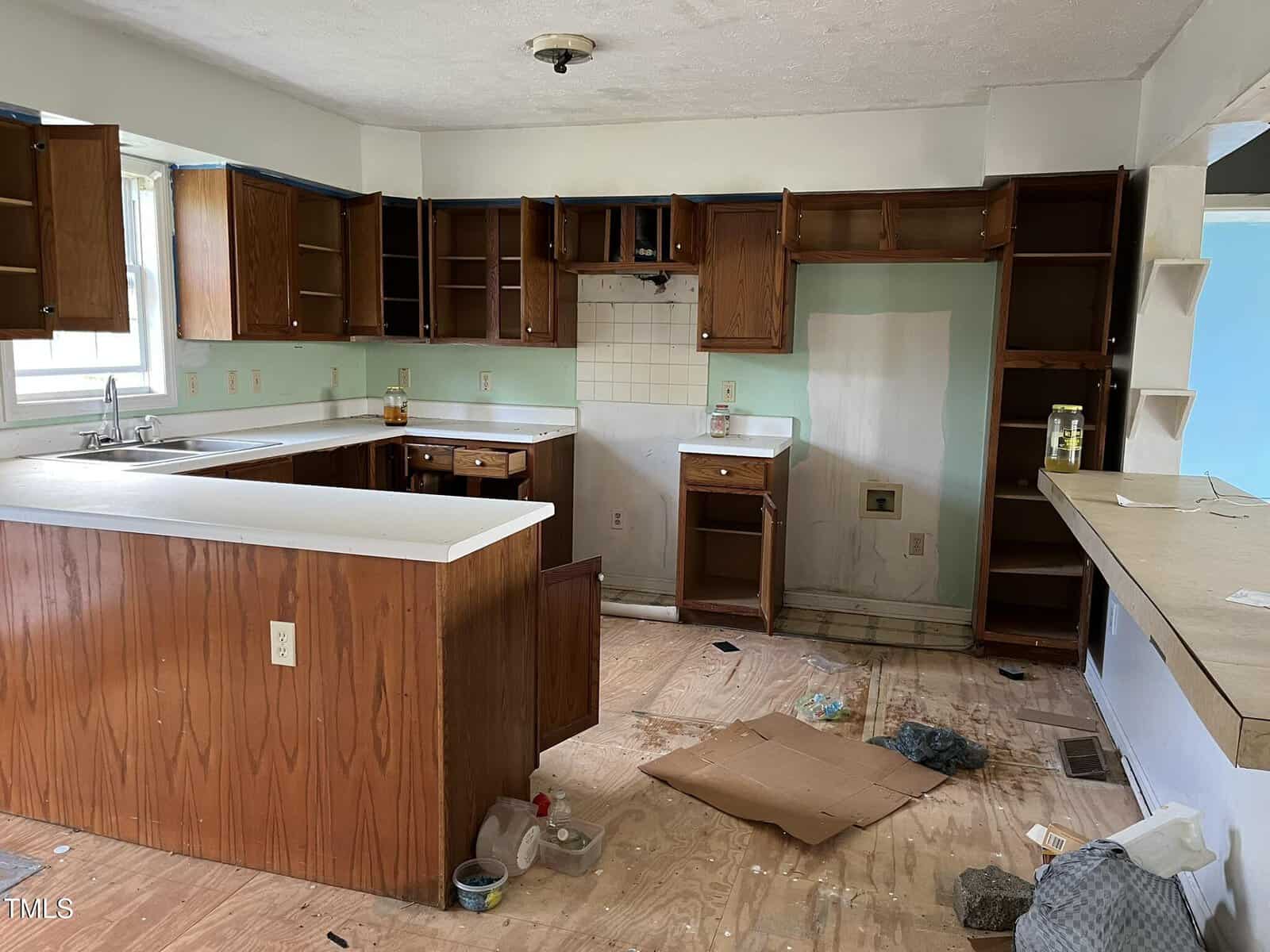 A vacant kitchen with unfinished floors, empty wooden cabinets, scattered debris, missing appliances, and a countertop with cleaning supplies and trash. The space appears to be under renovation or in disrepair.